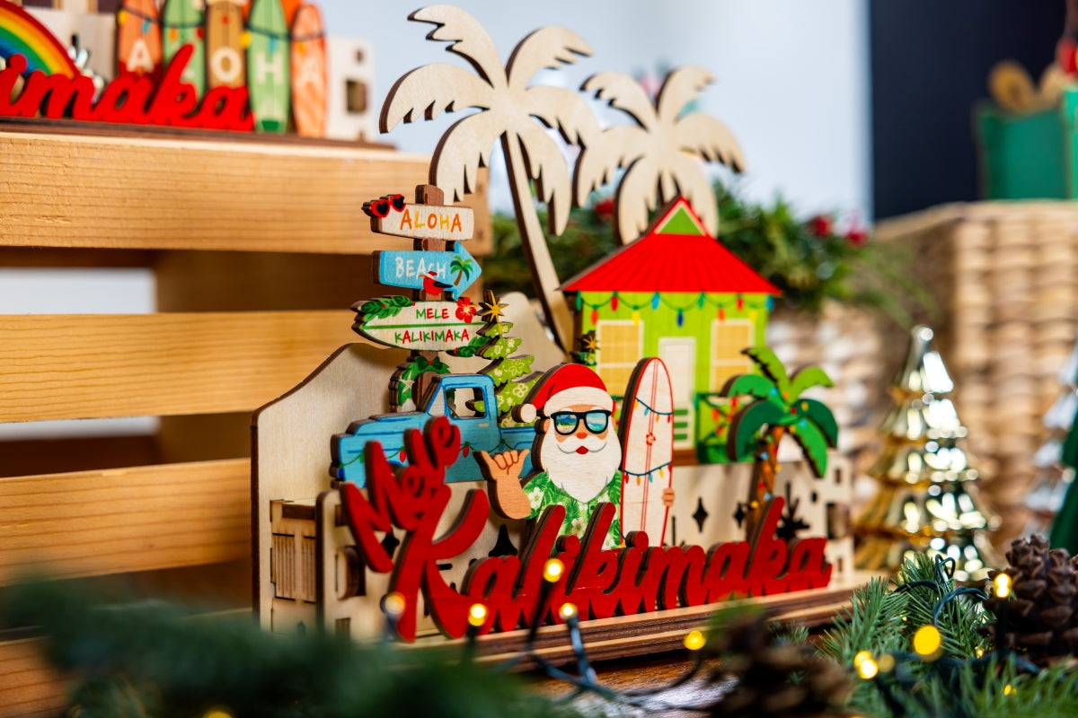Decorative 3D holiday tiered sign with Santa holding a surfboard, a blue truck, beach shack and other tropical elements with 'Mele Kalikimaka' script in a festive Christmas village scene