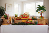 Decorative table setup with palm trees, baskets, and small figurines in a room with large windows.