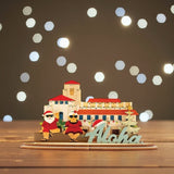 3D sign of Honolulu Hale building with Shaka Santa and Mrs Clause characters and 'Aloha' sign on a wood table with twinkling white lights in background