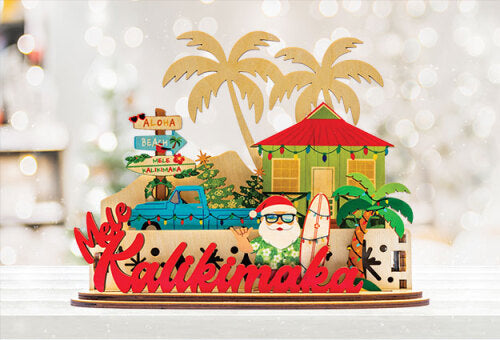Decorative 3D holiday tiered sign with Santa holding a surfboard, a blue truck, beach shack and other tropical elements with 'Mele Kalikimaka' script in front of white lights background